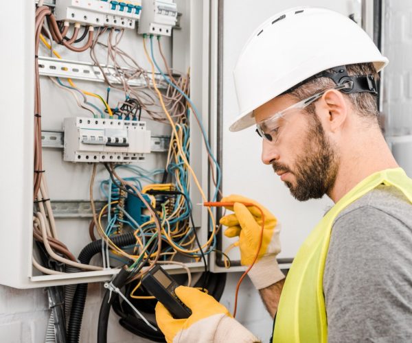handsome-electrician-checking-electrical-box-with-multimetr-in-corridor-e1624860078476